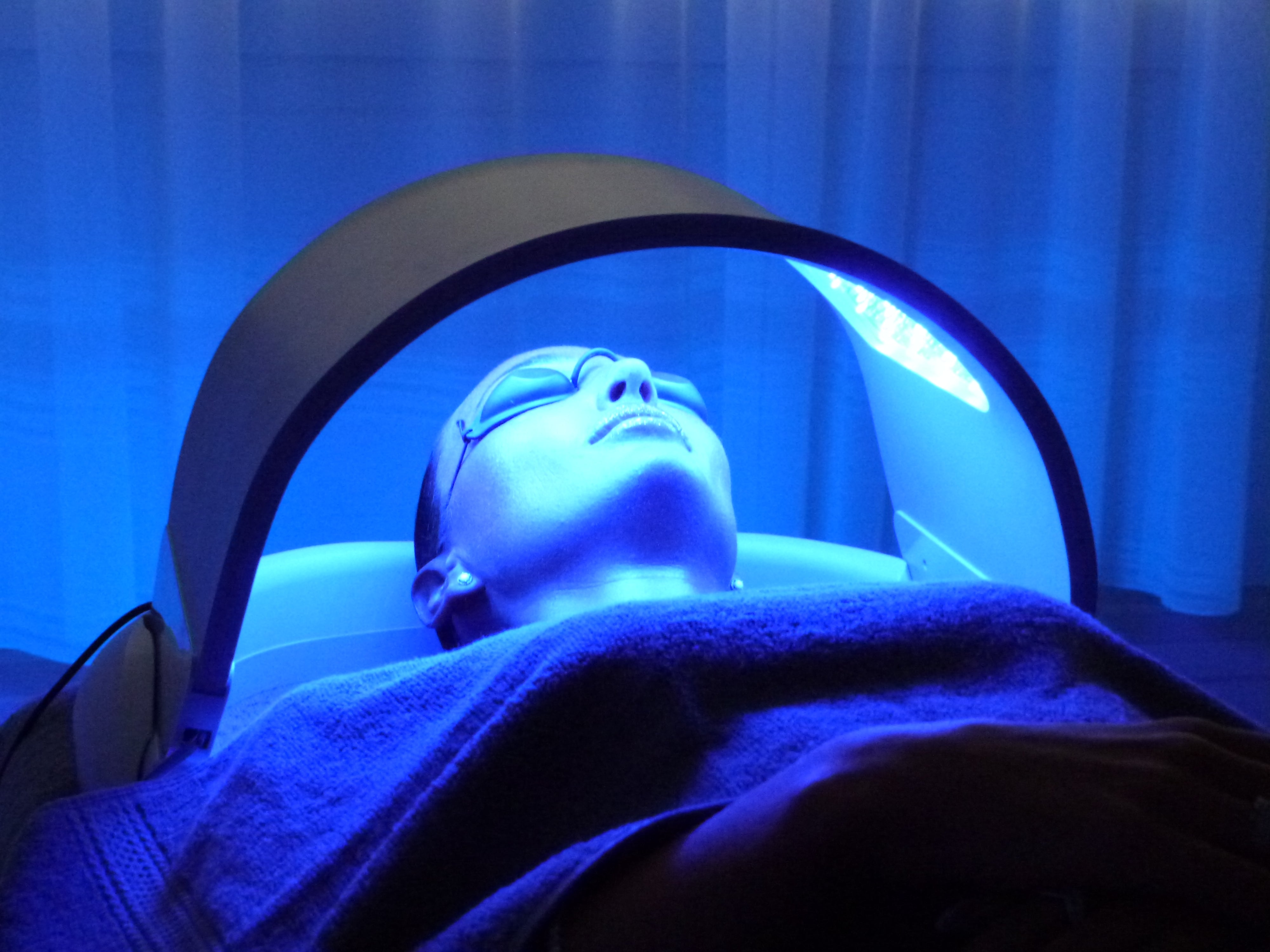 A person lying down under a curved blue light therapy device in a spa or treatment room, illuminated by a strong blue LED glow.