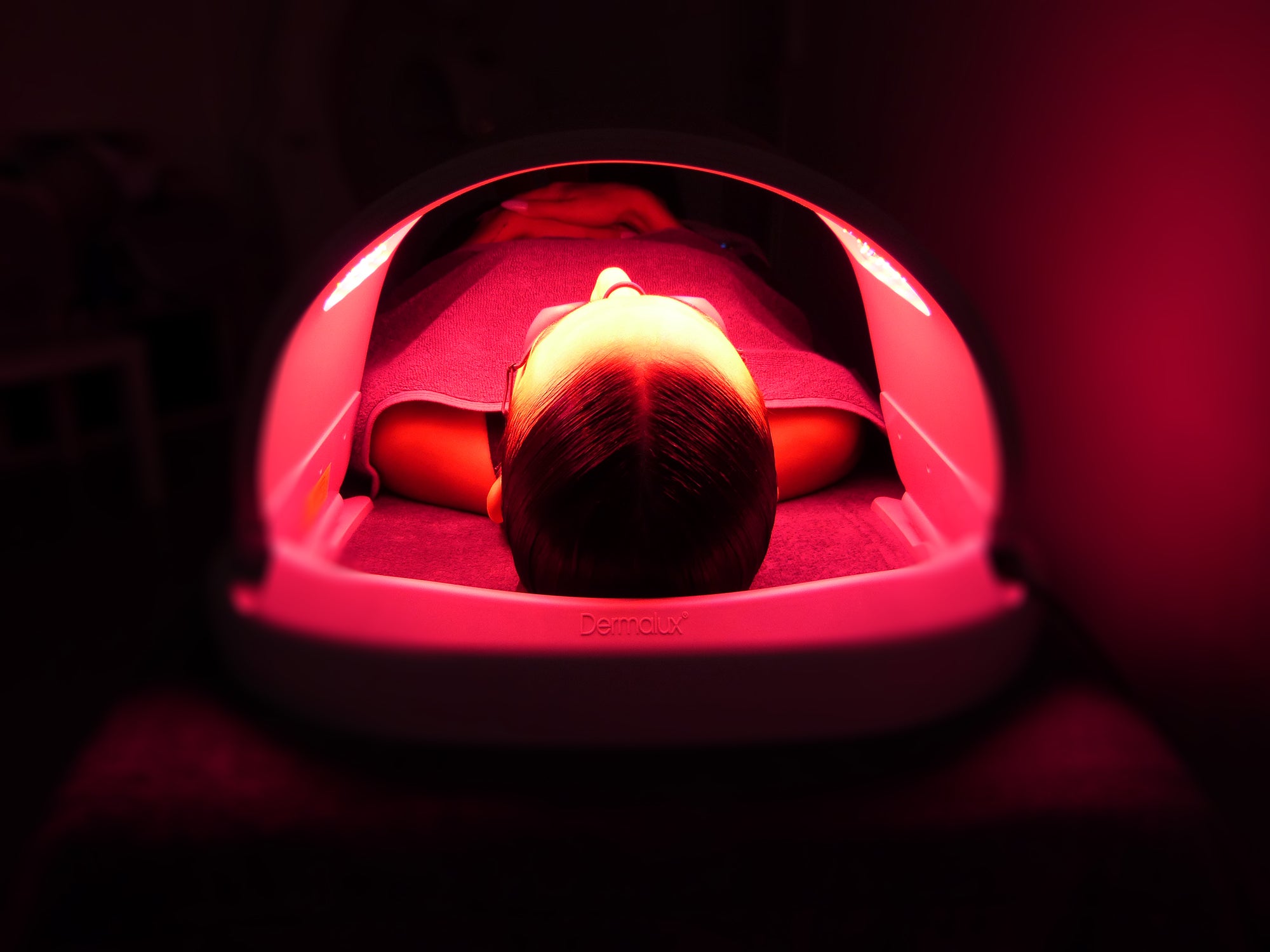 A person lying face down inside a red light therapy dome device, illuminated by a strong red glow in a dark room.